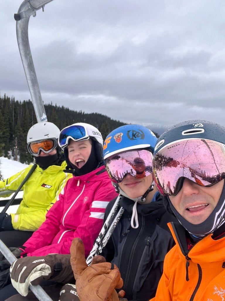 Cuatro personas con casco y gafas de esquí sonríen juntas en un telesilla, en un día nublado de montaña.