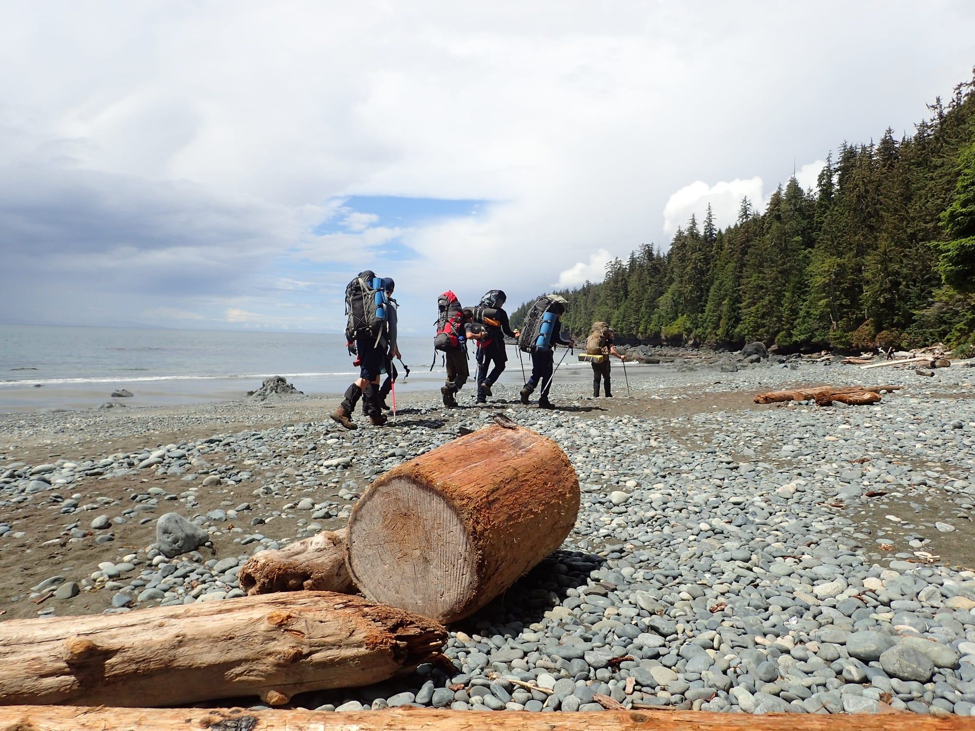 Grupo de adolescentes con mochilas de trekking camina por una playa de cantos rodados en la costa de Vancouver Island.