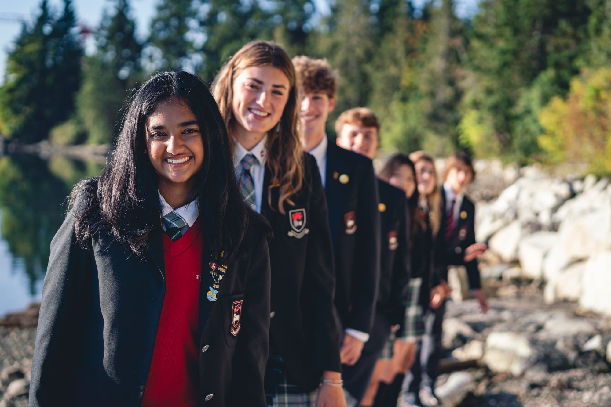 Grupo de estudiantes con el uniforme de su boarding school junto al agua y el bosque del campus.