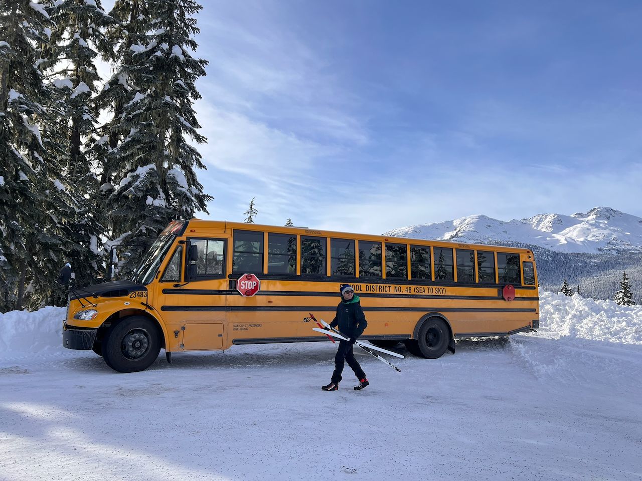 Un estudiante camina con sus esquís frente a un autobús escolar amarillo del distrito Sea to Sky, con montañas nevadas al fondo.