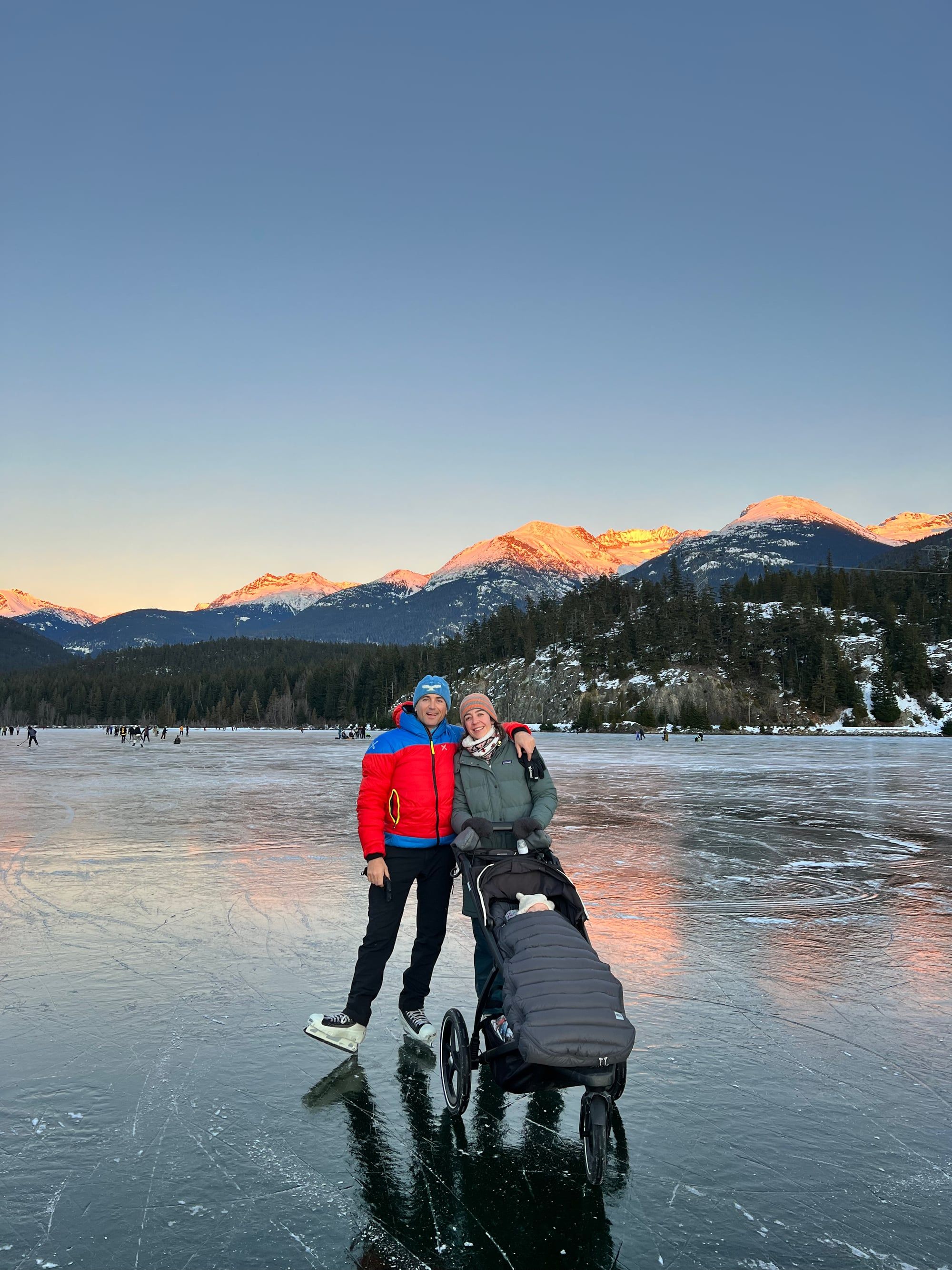Goretti y Adrià patinando sobre un lago helado al atardecer, con su bebé en el cochecito y las montañas iluminadas al fondo.